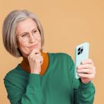 Oudere vrouw met bril kijkt bedachtzaam naar haar smartphone en houdt haar hand onder haar kin.