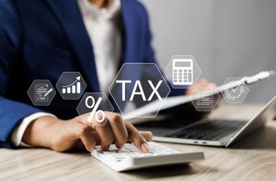 A professional in a blue suit calculating taxes using a calculator at a desk with a laptop and digital icons of documents, percentage sign, and tax symbol floating above the desk, representing online banking services. The person is focused on calculating taxes.