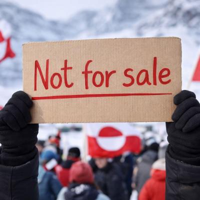 Een persoon houdt een kartonnen bord omhoog met de tekst “Not for sale”, terwijl op de achtergrond een menigte staat met rood‑witte vlaggen in een besneeuwd berglandschap.