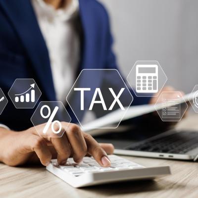 A professional in a blue suit calculating taxes using a calculator at a desk with a laptop and digital icons of documents, percentage sign, and tax symbol floating above the desk, representing online banking services. The person is focused on calculating taxes.