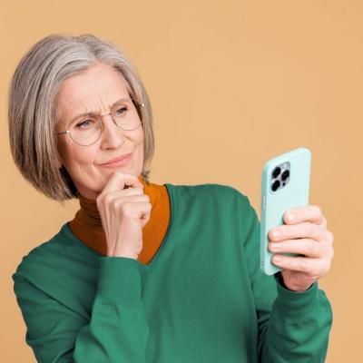 Oudere vrouw met bril kijkt bedachtzaam naar haar smartphone en houdt haar hand onder haar kin.