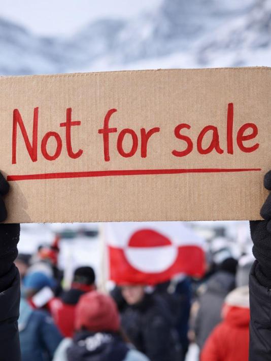 Een persoon houdt een kartonnen bord omhoog met de tekst “Not for sale”, terwijl op de achtergrond een menigte staat met rood‑witte vlaggen in een besneeuwd berglandschap.