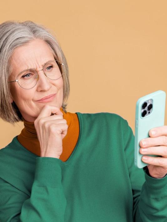 Oudere vrouw met bril kijkt bedachtzaam naar haar smartphone en houdt haar hand onder haar kin.