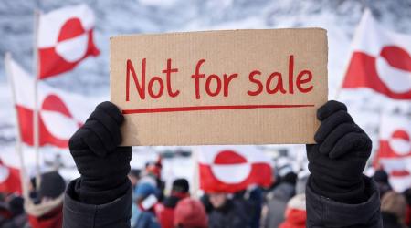 Een persoon houdt een kartonnen bord omhoog met de tekst “Not for sale”, terwijl op de achtergrond een menigte staat met rood‑witte vlaggen in een besneeuwd berglandschap.