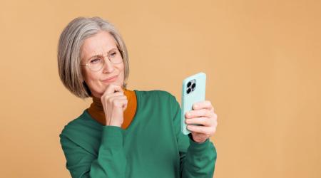 Oudere vrouw met bril kijkt bedachtzaam naar haar smartphone en houdt haar hand onder haar kin.